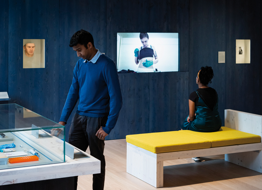 Wellcome Gallery, with one person sitting wathcing a screen with a person wearing prostehtic gloves. Another man is standing on the left looking into a glass case.
