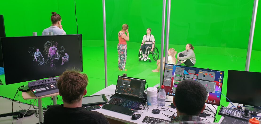 Two producers in a booth sit with multiple computer screens in front of them on a desk. On a green screen stage in front of them are 5 performers. 2 are standing, one is in a wheelchair and another two appear to be listening as they sit on the ground.