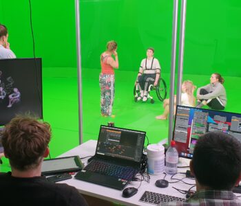 Two producers in a booth sit with multiple computer screens in front of them on a desk. On a green screen stage in front of them are 5 performers. 2 are standing, one is in a wheelchair and another two appear to be listening as they sit on the ground.