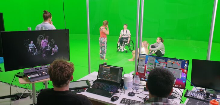 Two producers in a booth sit with multiple computer screens in front of them on a desk. On a green screen stage in front of them are 5 performers. 2 are standing, one is in a wheelchair and another two appear to be listening as they sit on the ground.